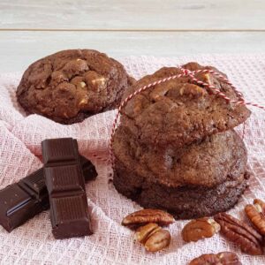 Chocoladekoekjes met pecannoten. Er zijn drie chocoladekoekjes op elkaar gestapeld en bijeen gebonden met rood-wit keukentouw. Om de koekjes heen liggen stukke pure chocolade en pecannoten.