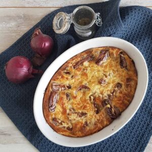 Gratin van rode ui in een kleine witte ovalen ovenschaal. Naast de schaal liggen enkele rode uien en een glazen potje met gedroogde tijm.