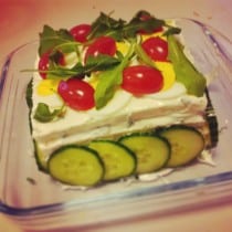 Een zijaanzicht van een Smorgastorte (Sandwichtaart), drie lagen brood met ertussen een smeersel van eiersalade. De taart is versierd met plakjes komkommer, tomaat en rucola.