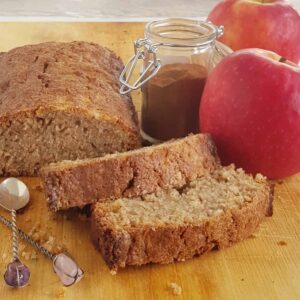 Appelcake op een houten serveerplank. Op de achtergrond een potje kaneel en twee appels.