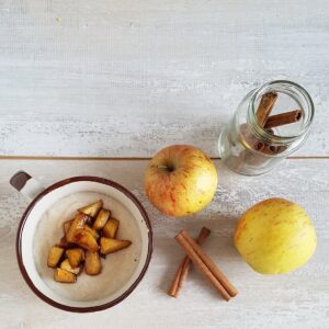 Een kom griesmeelpap met appel en kaneel, ernaast liggen staafjes kaneel en appels.