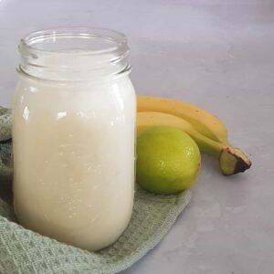 Smoothie met limoen en banaan in een grote glazen Mason Jar. De glazen pot staat op een lichtgroene doek, waar ook een banaan en een limoen op liggen.