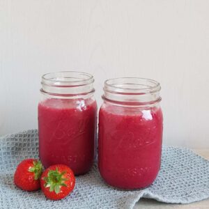 Smopothie met aardbei en rode biet in grote glazen Mason Jars. De smoothies staan op een blauwe doek waar ook wat verse aardbeien op liggen.