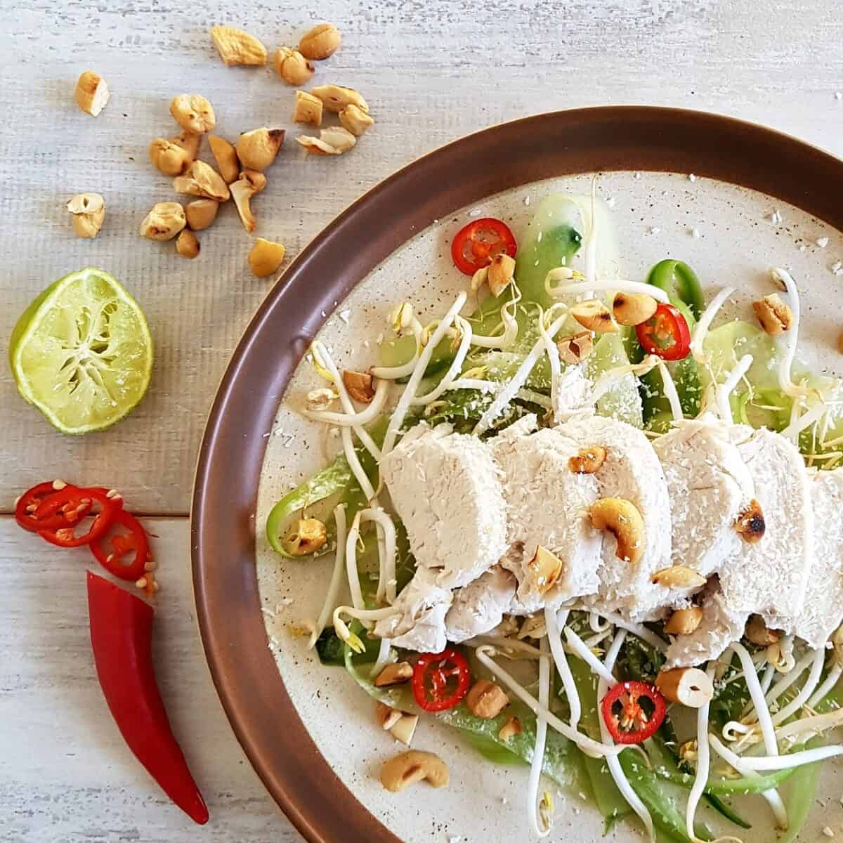 Gepocheerde kokoskip met komkommersalade op een bord, met limoen, rode peper en cashewnoten als garnering.