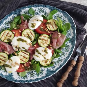 Salade Caprese - Maaltijdsalade met tomaat, mozzarella, basilicum, pesto, gegrilde courgette en rauwe ham, op een ouderwets bordje.