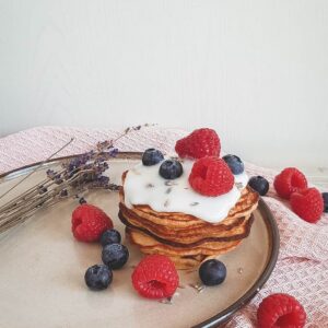 Een stapel boekweit pannenkoeken op een bruin bord, gegarneerd met een schep yoghurt. Om de pannenkoekjes heen ligt vers fruit (frambozen en blauwe bessen).