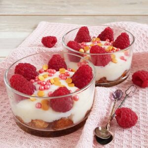 Trifle met custard en frambozen in kleine glazen bakjes. Er zijn feestelijke sprinkles over de trifles gestrooid.