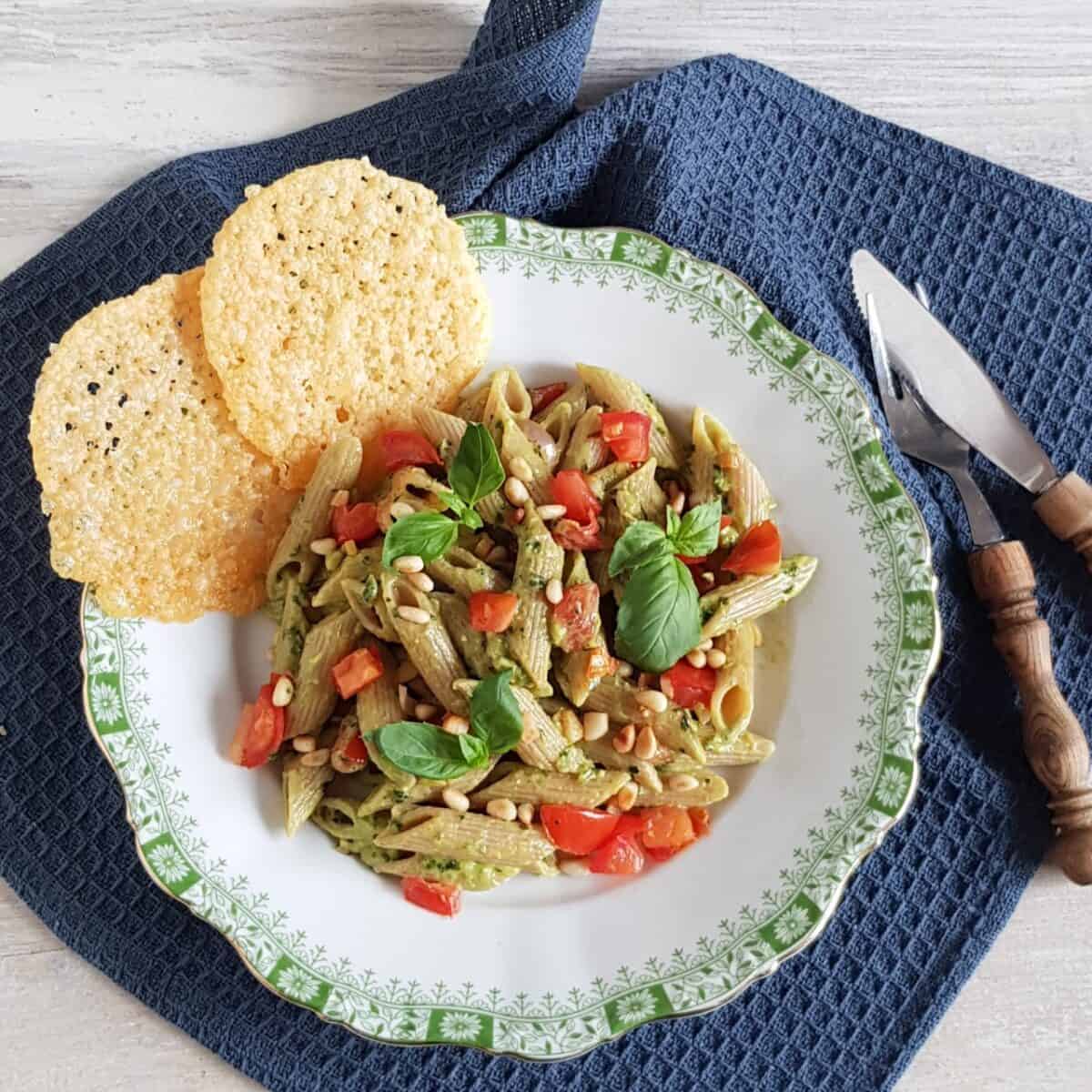 Pasta met avocadosaus, tomaat en pijnboompitten. Naast de avocadopasta liggen krokant gebakken parmezaankoekjes uit de oven.