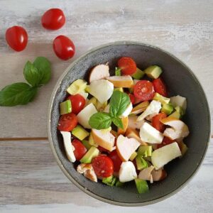 Salade met gerookte kip, avocado en mozzarella in een kom. Ernaast wat tomaatjes en basilicum ter decoratie.