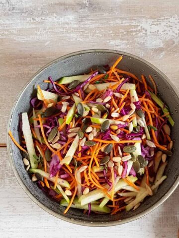 Rode koolsalade met appel en pittenmix, in een grijze kom met bestek ernaast.