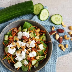 Spinaziesalade met geitenkaas, geraspte wortel en noten, in een grijze kom die op een blauwe doek staat.
