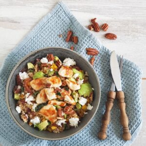 Rijstsalade met kip, avocado en feta, gegarneerd met pecannoten. De salade zit in een grijze kom en staat op een blauw doekje.