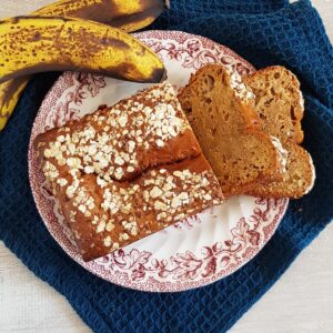 Bananenbrood met havermout-topping op een ouderwets bordje. Naast het bord ligt een overrijpe banaan.