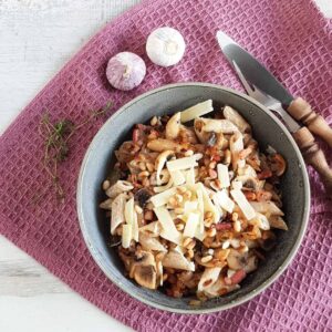 Pasta met spek, champignons en kruidenkaas in een grijze kom. De pasta is bestrooid met parmezaanse kaas en pijnboompitten.