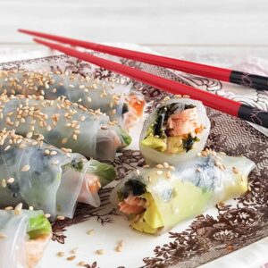 Springrolls met zalm en avocado op een bord. Eén van de springrolls is opengesneden zodat je de vulling van zalm, avocado en zeewier goed kunt zien. De rolletjes zijn bestrooid met sesamzaad.