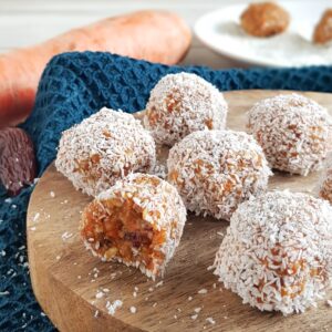 Carrot cake balletjes op een houten serveerplank. De kleine tussendoortjes zijn gevuld met wortel en dadel, en zijn in kokosrasp gerold.