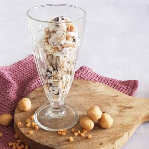Chocolate chip cookie dough ijs in een hoog milkshakeglas. Het glas met ijs staat op een houten plank en er liggen mini stukjes fudge en cookie dough balletjes omheen.