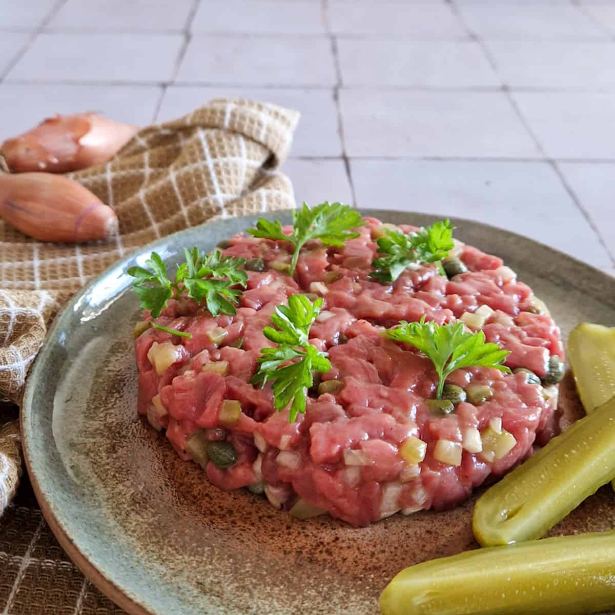 Steak tartaar als voorgerecht, geserveerd op een bordje met stukken augurk ernaast.