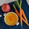 Wortelsoep met appel en gember, de soep is gegarneerd met sliertjes wortel en er liggen verse wortels naast de kom soep.