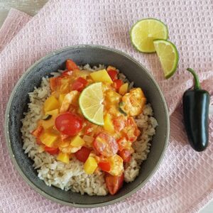 Garnalencurry met kokosmelk in een grijze kom op een roze doek. De curry zit vol verse groenten en is gegarneerd met limoen.