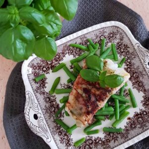 Kabeljauw met pesto en parmaham op een stapeltje sperziebonen. Er staat een basilicumplant naast het bord met vis.