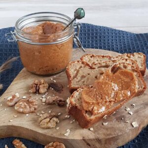 Notenspread in een potje die op een houten plank staat. Op de plank liggen plakken bananenbrood die zijn besmeerd met de spread, en er liggen walnoten bij.