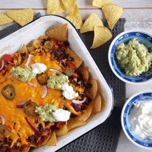 Nachos met gehakt en zure room in een ovenschaal. De nacho schotel is voorzien van een dikke laag geraspte cheddar en bestrooid met jalapenos, rode ui, guacemole en zure room.