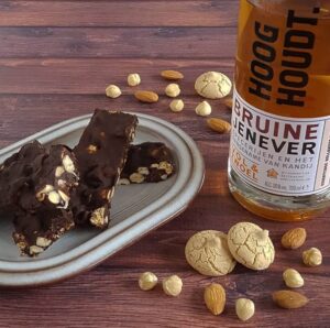 Een schaaltje met brokken chocolade met noten, rozijnen en jenever. Er omheen liggen verschillende soorten noten en koekjes, de ingrediënten van dit recept.