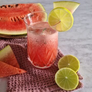Watermeloen cocktail in een cocktailglas, op een blauwe doek. Ernaast liggen een stuk watermeloen en een gehalveerde limoen.