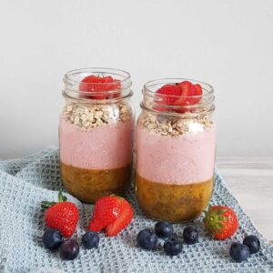 Chiapudding met smoothietopping, havermout en vers fruit, in grote glazen Mason Jars die op een lichtblauwe doek staan. Voor de twee glazen ligt vers fruit (aardbeien en blauwe bessen).