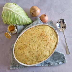 Spitskool schotel, gegratineerd met kaas. De ovenschotel staat op een blauwe doek en er omheen liggen enkele ingrediënten van het recept: uien, spitskool en kerrie.