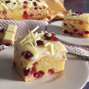 Cranberrycake: een close-up van een stuk cake met cranberries, een laag witte frosting en gedroogde cranberries. Op de achtergrond staat de rest van de taart.