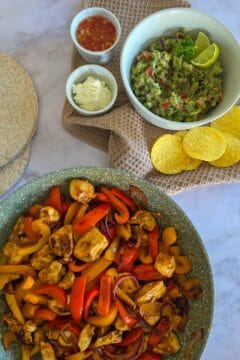 Een pan kip fajitas met ernaast volkoren tortilla's, een schaal guacemole en kleine schaaltjes met zure room en chilisaus.
