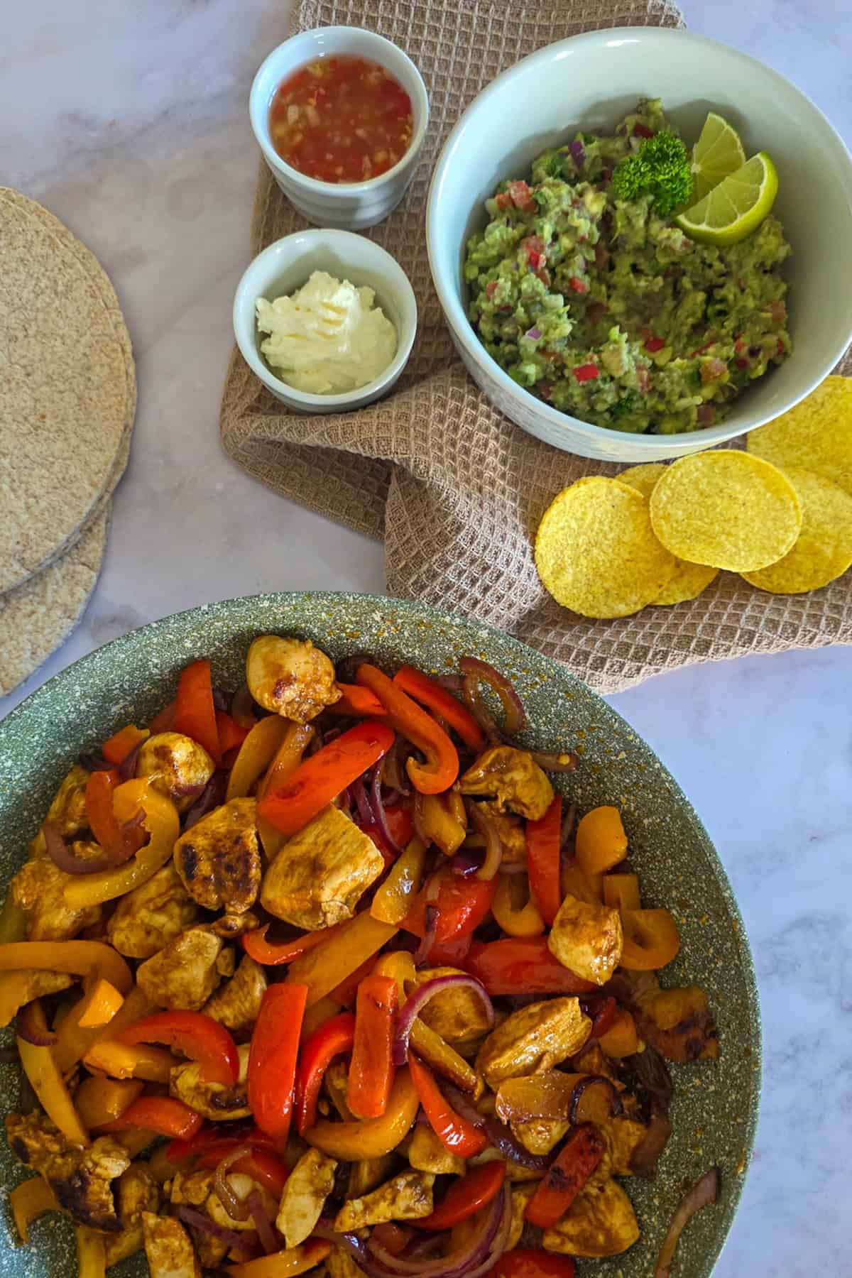 Een pan kip fajitas met ernaast volkoren tortilla's, een schaal guacemole en kleine schaaltjes met zure room en chilisaus.