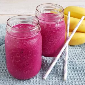 Smoothie rode biet banaan in grote Mason Jars met rietjes en bananen ernaast. De smoothies staan op een blauwe doek.