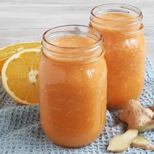 Sapje met wortel en gember in twee grote glazen Mason Jars. De glazen staan op een lichtblauwe doek en er ligt verse gember en sinaasappel omheen.