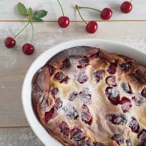 Een ovenschaal met kersenclafoutis, er liggen verse kersen omheen en de clafoutis is bestrooid met poedersuiker.