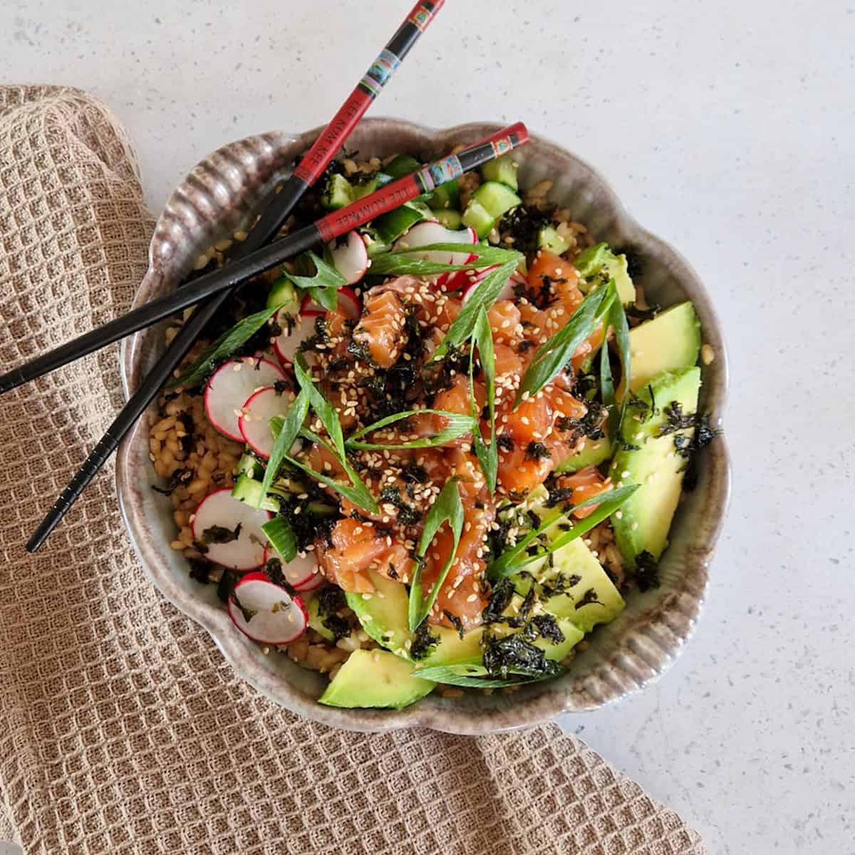 Poké bowl met zalm, avocado en diverse groenten. De bowl is gegarneerd met fijngesneden bosui en er liggen eetstokjes bij.