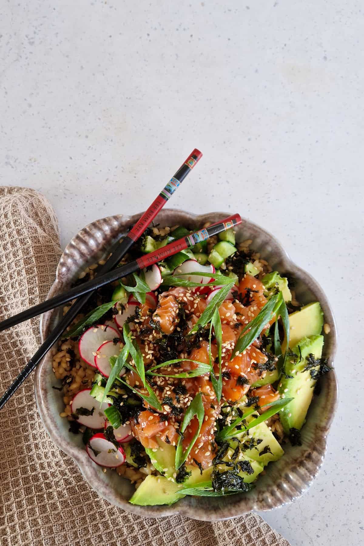 Een zalm poké bowl met diverse groenten en bosui als garnering. De kom staat op een beige doek.