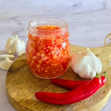 Een potje zelfgemaakte sweet chilisaus. De pot staat op een houten plankje met er omheen verse rode pepers en bollen knoflook.