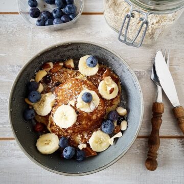 Een kom gevuld met een stapel havermout pannenkoeken, gegarneerd met plakjes banaan en blauwe bessen. Eromheen bestek, blauwe bessen en een pot met havermout.