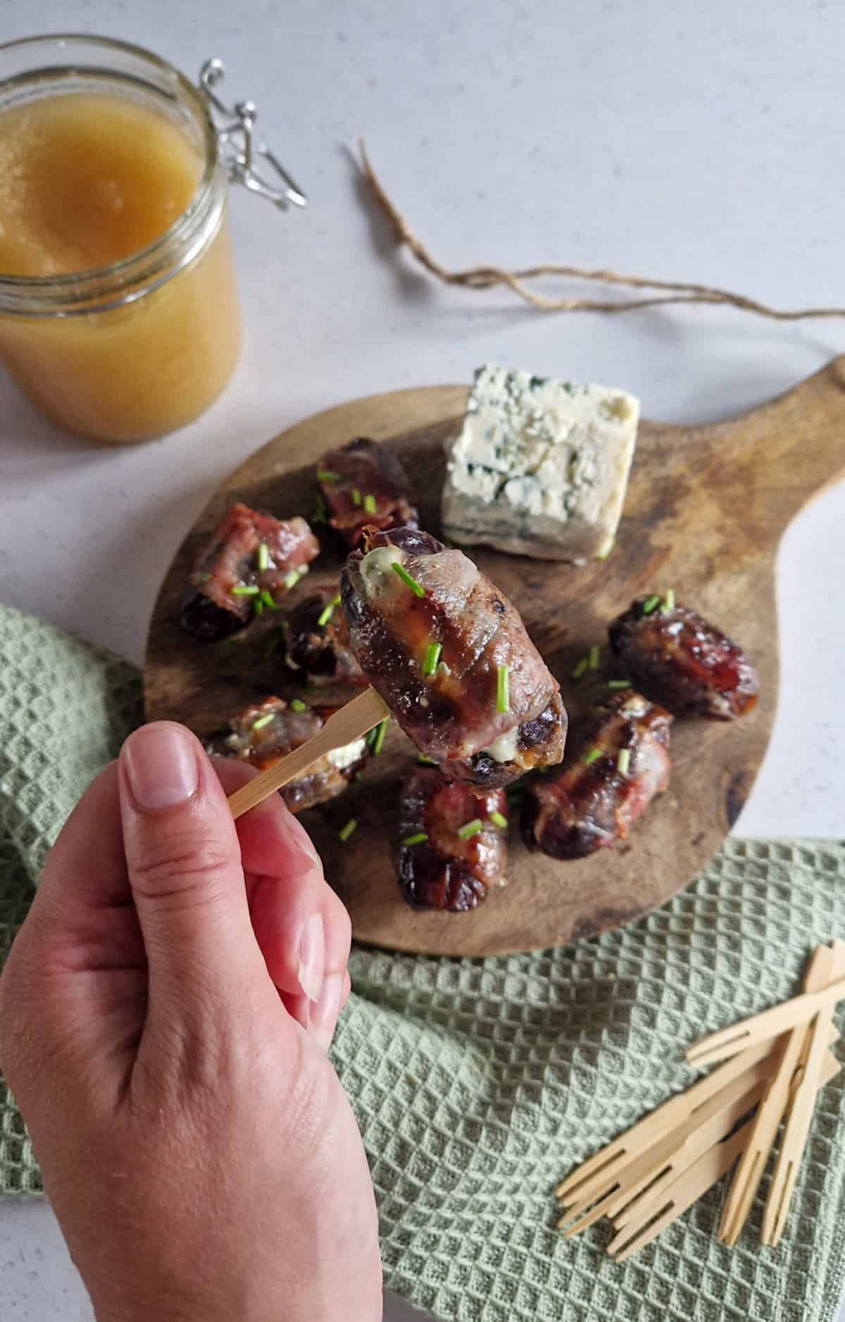 Dadels met blauwe kaas en spek op een borrelplank. Een hand pakt één van de hapjes met een prikker.