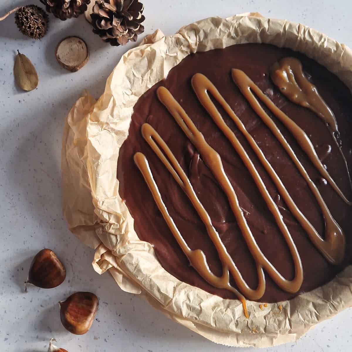 Een kastanje chocoladetaart in een bakvorm met bakpapier. Enkele kastanjes liggen er ter decoratie naast.