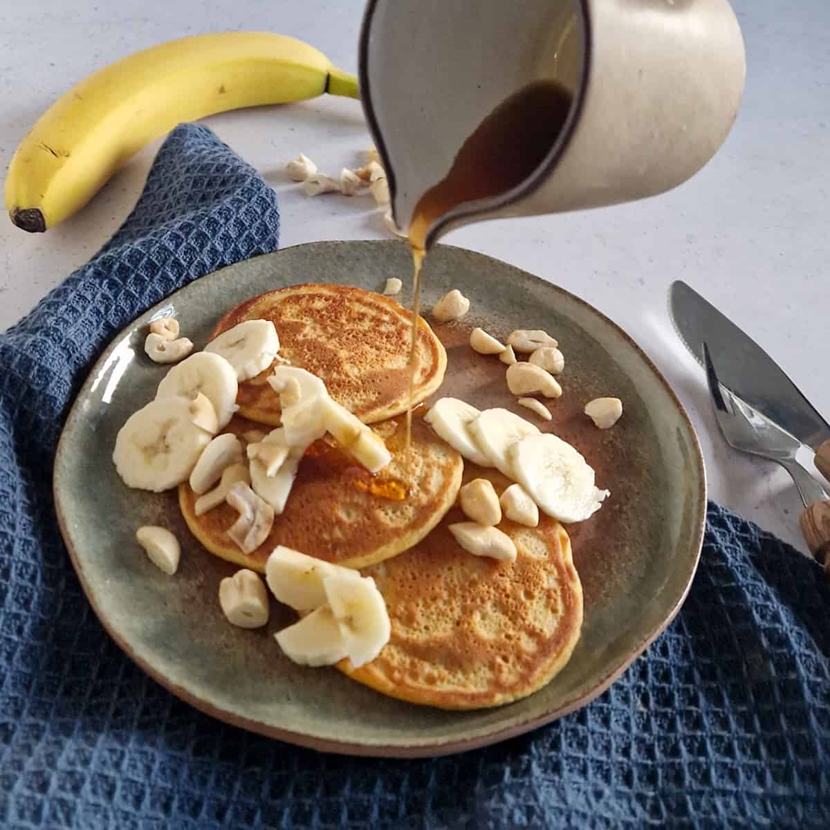 Een bord met zoete aardappelpancakes, gegarneerd met banaan. Er wordt maple syrup overheen gegoten uit een kannetje.