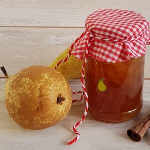 Perenjam in een glazen pot met rode doekje. Het potje is dichtgebonden met een rood-wit lintje en er ligt een verse peer en een stokje kaneel naast de perenjam.