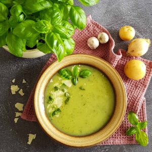 Courgettesoep, gegarneerd met flinters parmezaan en verse basilium. Naast het bord met courgettesoep staan een basilicumplant, en wat ingrediënten van het soep recept: knoflook, ui en aardappel.