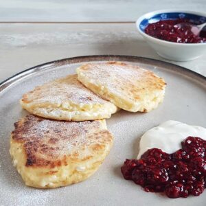 Cottage cheese pannenkoeken met crème fraiche en een compote van rood fruit. De pannenkoeken zijn bestrooid met poedersuiker.