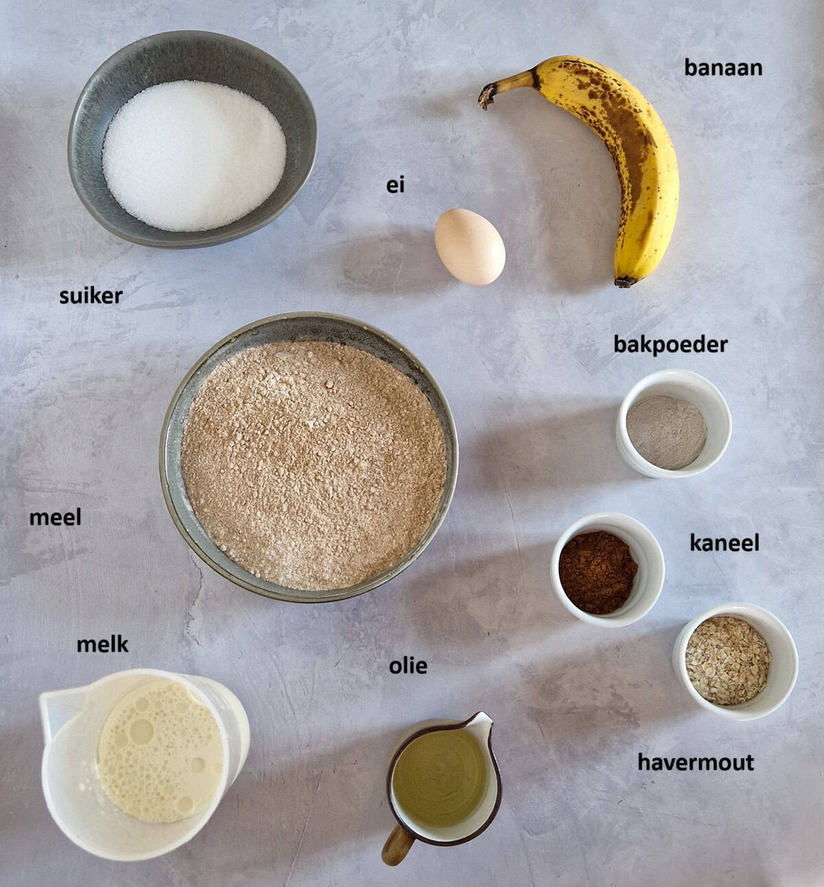 De ingrediënten om ontbijt muffins met banaan te maken, uitgestald op een lichtgrijze ondergrond.