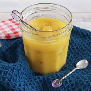 Passievruchten Curd in een glazen pot. Er staat een zilveren lepeltje in de pot en de pot staat op een blauwe doek.