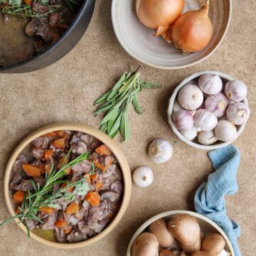 Een kom boeuf bourguignon met er om heen wat uien, knoflook en kruiden.